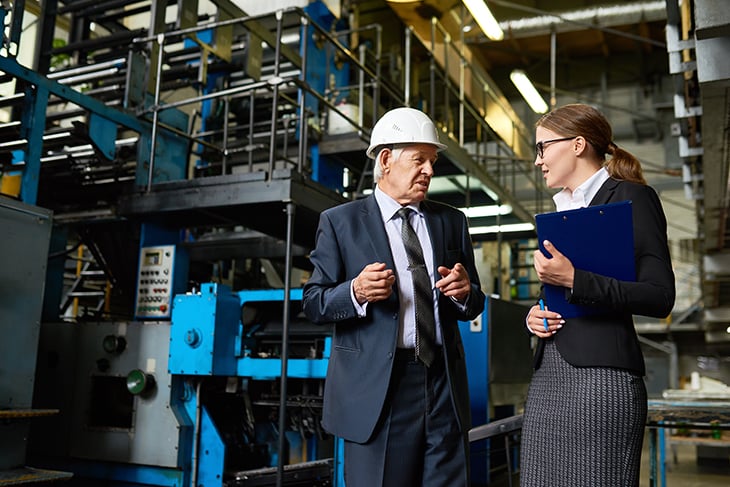 Businesswoman and older businessman in a hard hat having a conversation inside a factory.