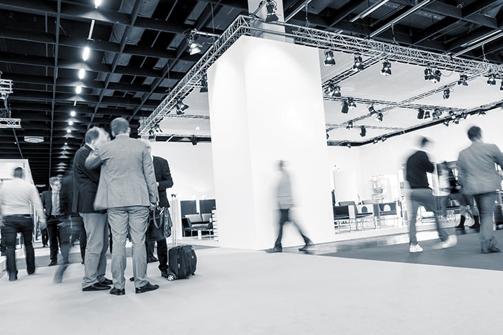 Black-and-white, low-angle photo of attendees walking the floor of a trade show in a large convention center.