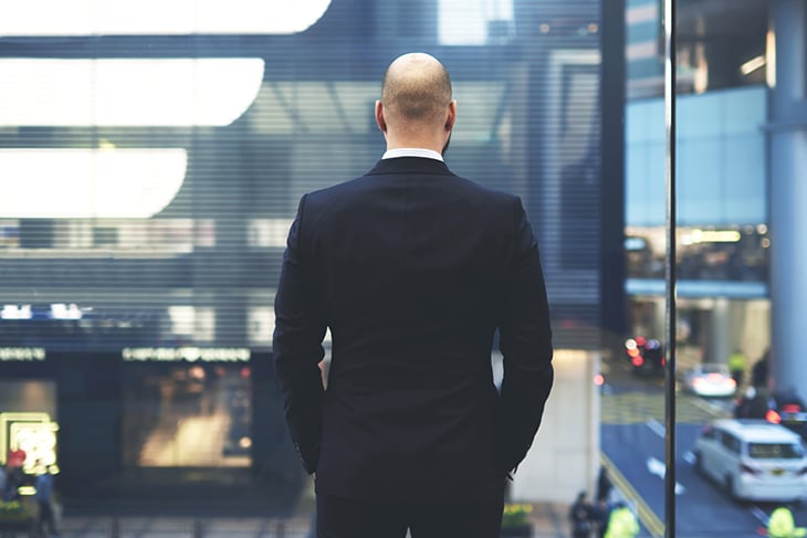 Balding male, wearing a suit, staring out large floor to ceiling windows